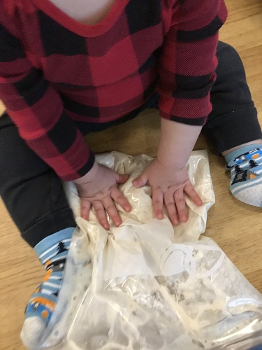 A child sitting on the ground squishing a ziplock bag filed with ingredients to make dough