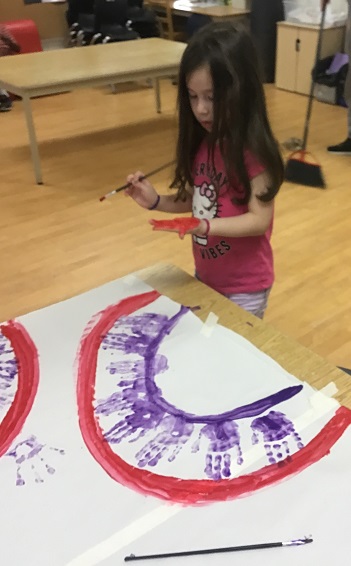 child standing in front the beginning of a rainbow painting. she is painting her hand red to add above the red arch of the rainbow. 