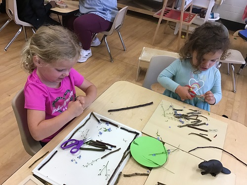 2 girls sitting at a table creating some art with small twigs and sticks