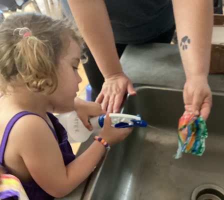 child standing at a sink holding a spray bottle and spraying a rainbow, held by an educator, that she coloured.