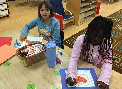 two children sitting at a table with a basket full of markers. The children are colouring.