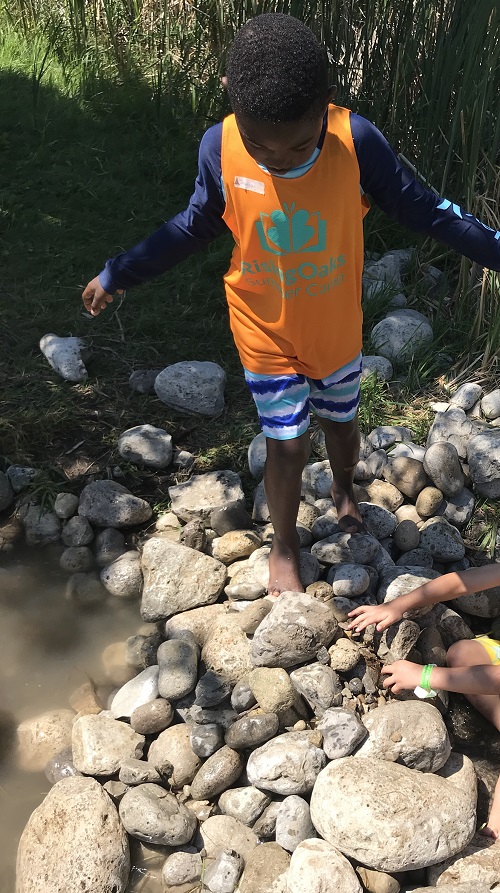 child in orange camp water tank top walking along rocks near a water stream,
