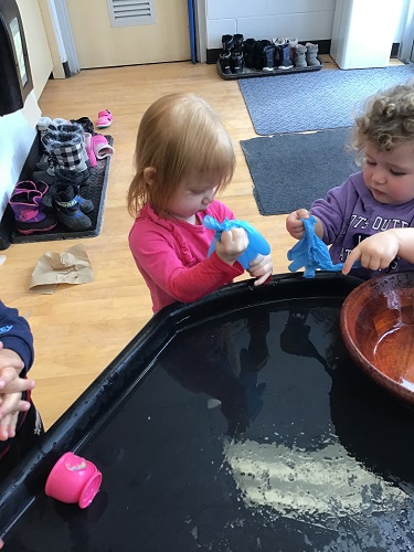 2 toddler children standing at the sensory table holding and looking at gloves they are holding in their hand