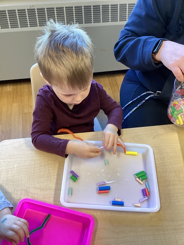 A child at a table stringing small pieces of colourful straws onto a string