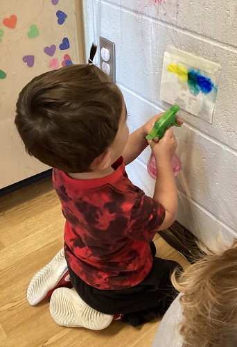 A child sitting on the floor spraying water onto a coloured piece of paper taped to the wall