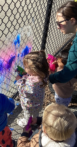Children standing at a fence spraying paint onto a clear piece of plexiglass
