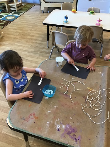 Two children sitting at a table putting glue and string on a piece of paper