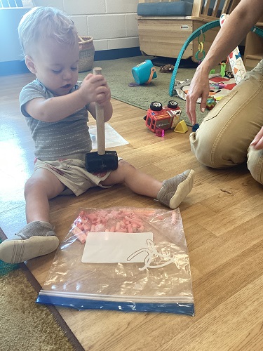 A child sitting on the ground using a mallet to crush chalk in a ziploc bag
