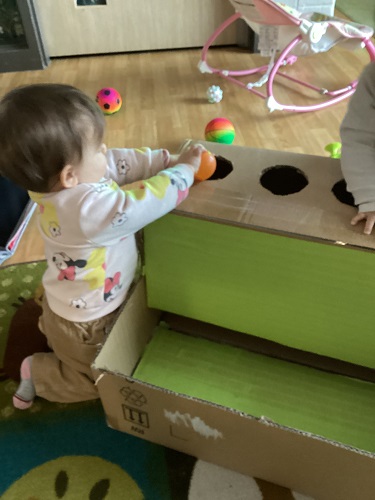 An infant kneeling beside a cardboard ball drop