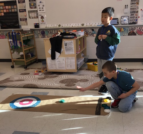 Two children in a classroom, practicing curling on the classroom floor