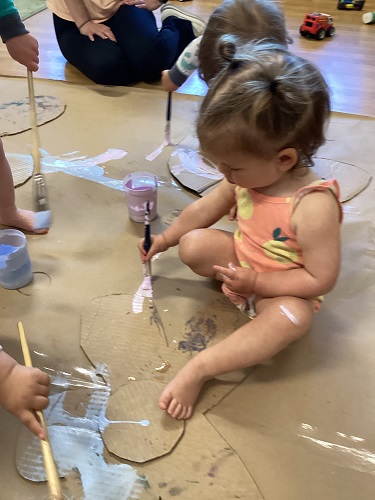 An infant painting on a cardboard flower on the floor