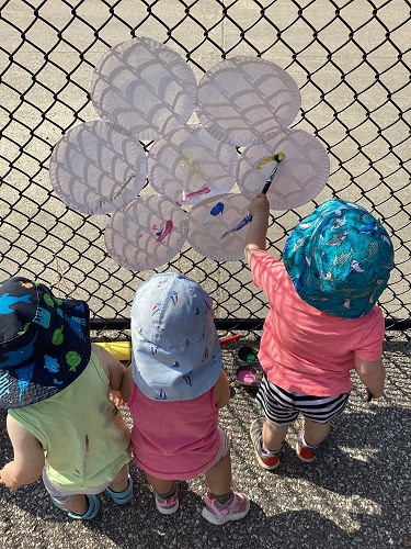 Children outside at the fence painting a paper plate flower