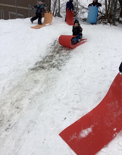 1 School Age child sitting on their bum on a red sled going down the hill with a smile on his face, another School Age child walking up the hill holding a sled and 2 other SChool Age 1 children standing at the top of the hill holding onto their sleds waiting their turn to go down the hill