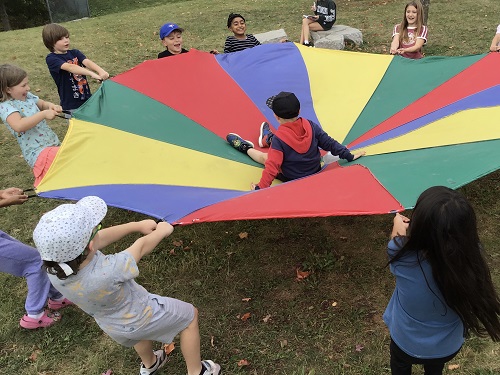 8 School age children holding and pulling back on the sides of the parachute, one school age child sitting in the middle of the parachute