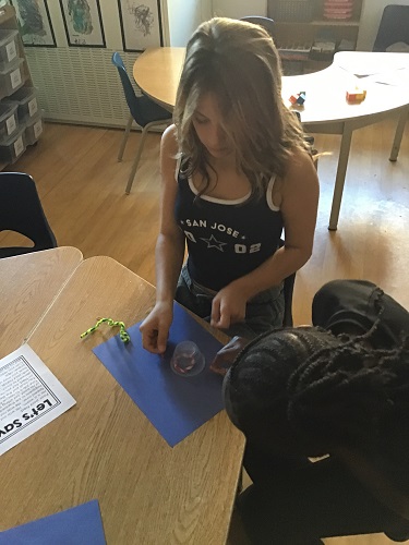2 school age children sitting at the table looking at a cup with pipe cleaner under the cup, and paper clips in their hands