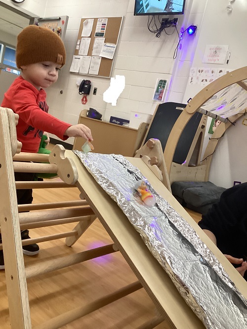 A toddler child putting little people figures down a ramp covered in tin foil