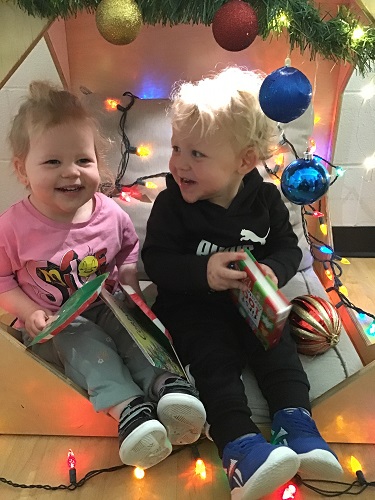 2 Preschool children sitting in the book cube with books on their lap, one child smiling at an educator, the other child smiling at the other child