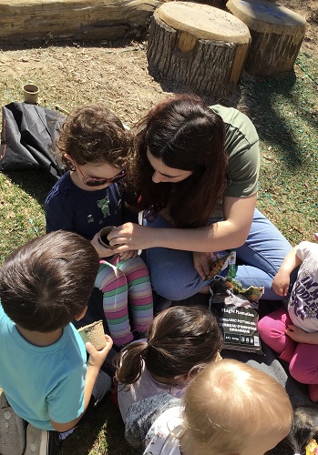 5 Toddlers and a toddler educator, one toddler is holding a cup with soil as the educator is putting something in the cup