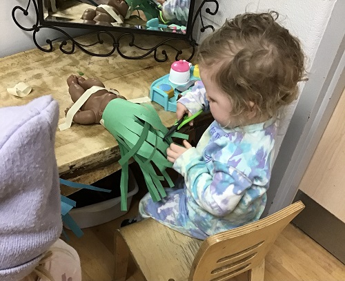A Preschool child sitting at a table with a baby doll and construction paper with cut slits, the child has scissors and is cutting off the paper hair
