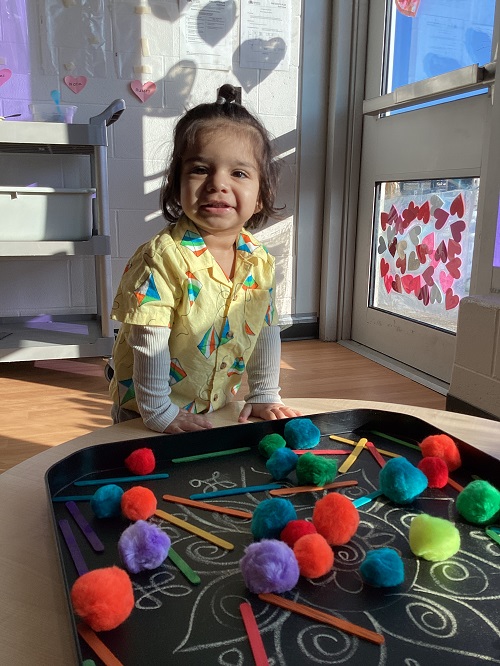 An infant child at a tuff tray with colourful pom poms and popsicle sticks