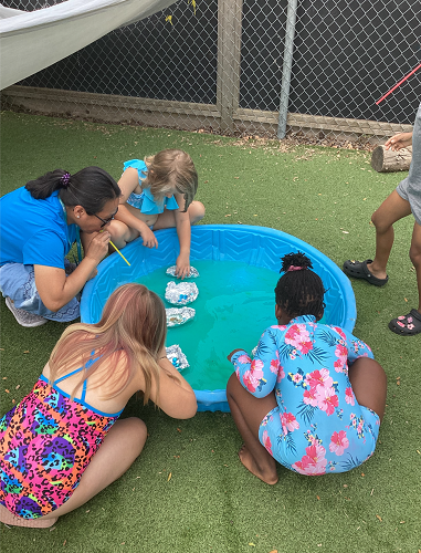 3 summer camp children sitting on the outside of a kiddie pool with tin foil boats floating in the pool, the camp children are using straws to move the boats and an educator has joined them
