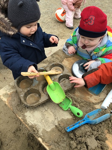 Two toddlers are making sand cakes on the playground.