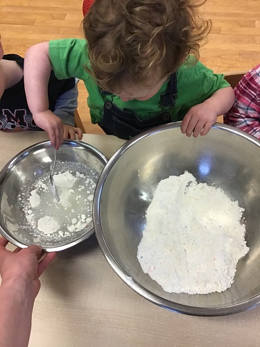 A toddler is adding dry ingredients to wet ingredients.