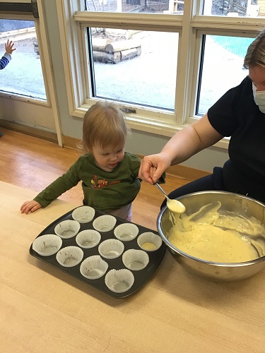 A toddler is watching an educator spoon cupcake batter into muffin pan.