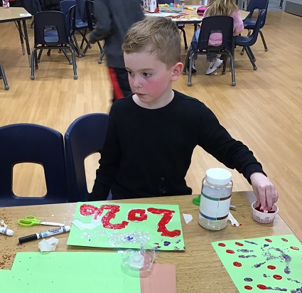 A school age child sitting at a table using paint and glitter to create a 2026 poster