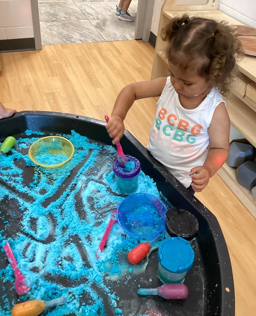 A child using various materials to explore with coloured salt in a tuff tray.