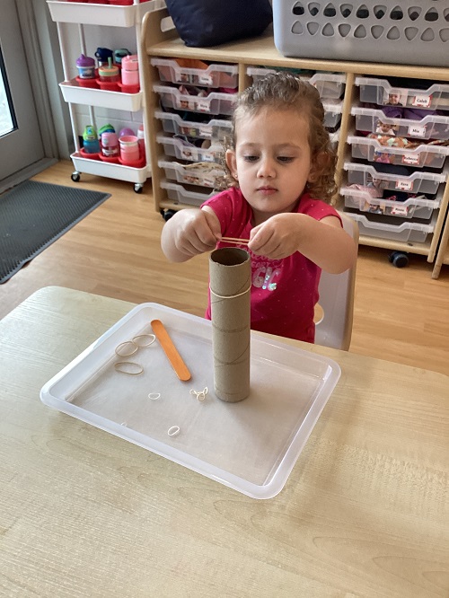 A child maneuvering an elastic band around a paper towel roll.