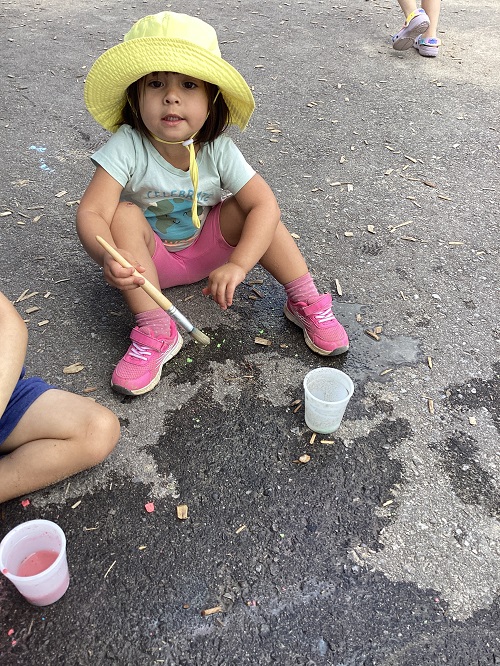 A child sitting on the ground outside while holding a paintbrush. 