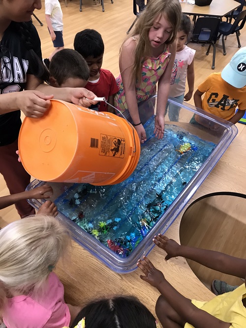 A group of children observe as a bucket of water is poured into the sensory bin.