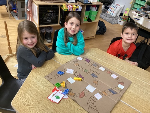 3 School Age children sitting at the table with the board game in front of them all looking at the camera and smiling