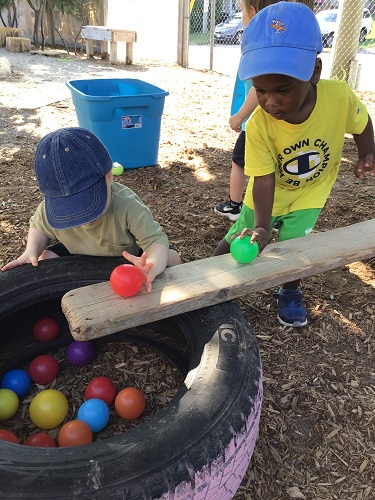 2 Toddler children using a balance beam to place balls onto and watch them roll down the ramp