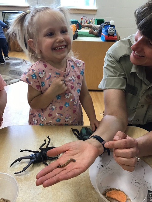 Toddler standing with a volunteer from the butterfly conservatory smiling big, there is a larva in the volunteers hand