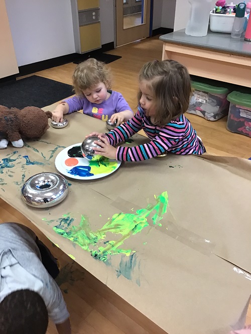 2 Toddler children at the table - one Toddler holding two m irror balls and another Toddler dipping the mirror ball in paint on a plate