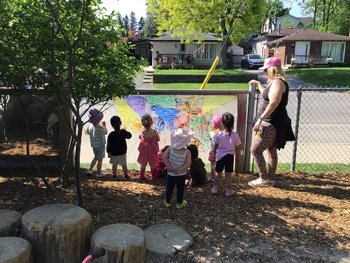 8 children standing looking at the window with paint while an educator watches the children