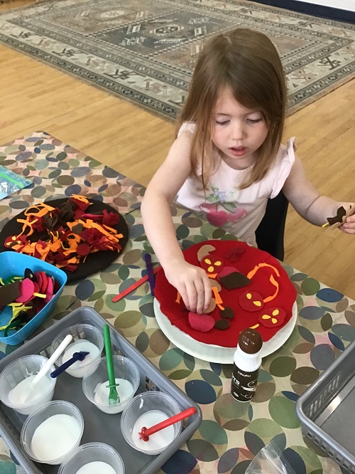 School Age child sitting at that able with a felt pizza and placing felt toppings on top of the pizza