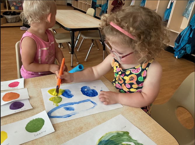2 Toddler children sitting at the table, one looking away, the other one sitting with a paint brush, paint and paper
