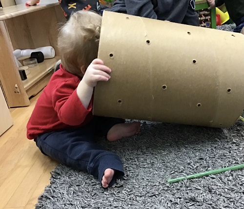 An infant is holding up a cardboard tube and looking into is by sticking their entire face into the tube.