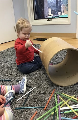 An infant is putting a straw into a small hole in a cardboard tube.