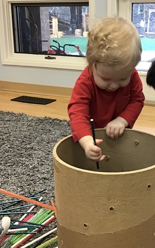 An infant is trying to put a straw through a hole in a cardboard tube.