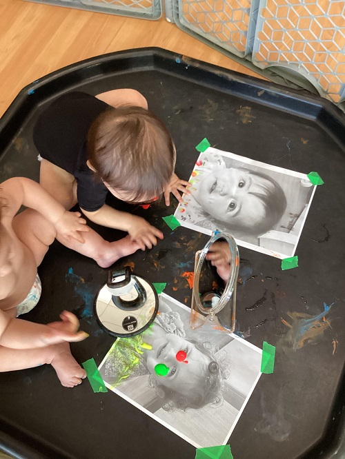 2 infants sitting on a tuff tray with mirrors and photos of themselves and finger paint