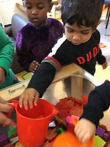 A toddler child filling a red measuring cup with leaves while another child looks on from behind. 