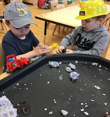 2 Preschool children sitting at the tuff tray with clay, trucks, and bugs - one child wearing a construction hat
